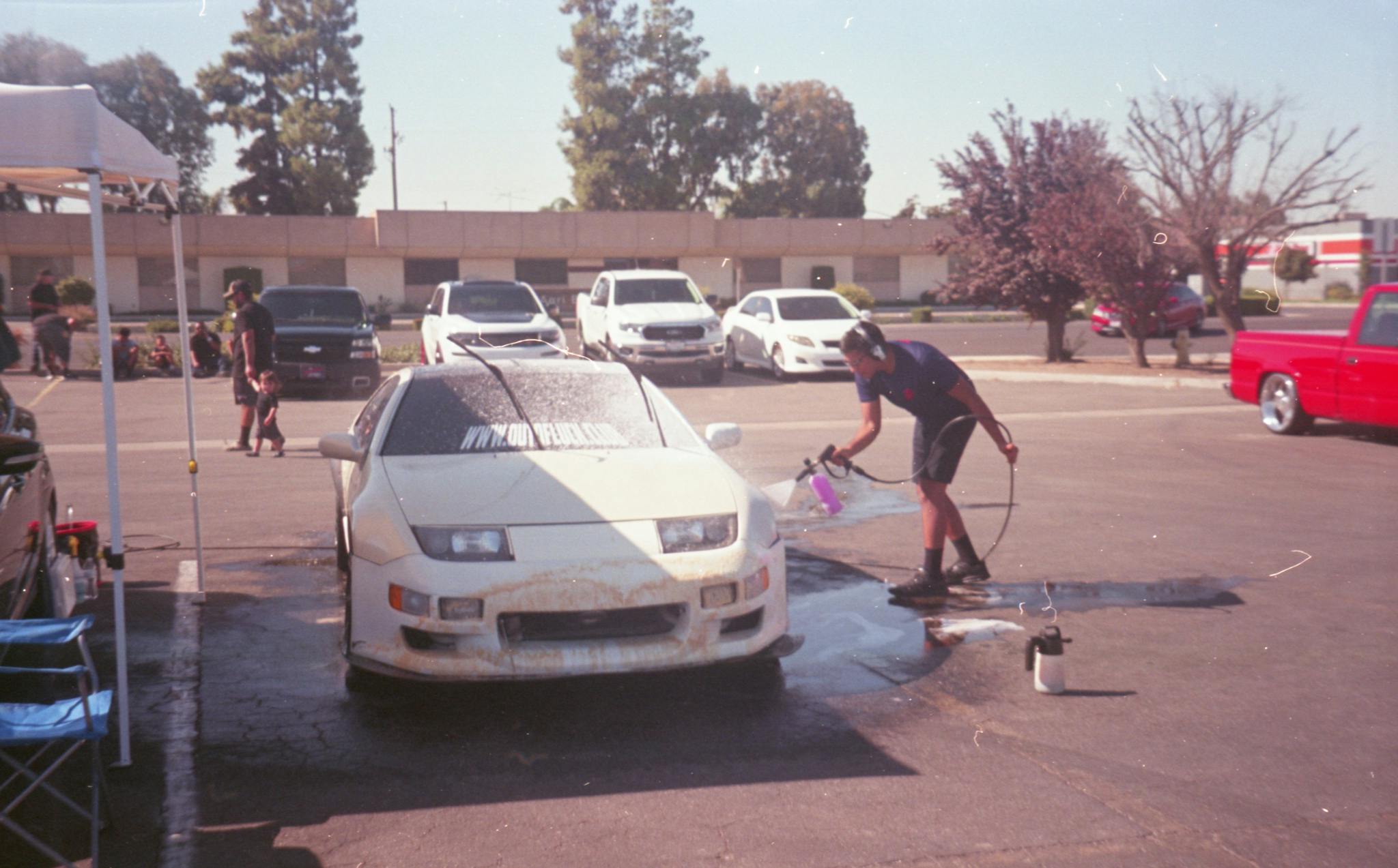 A man using a hose to wash a car in an outdoor parking lot on a sunny day.