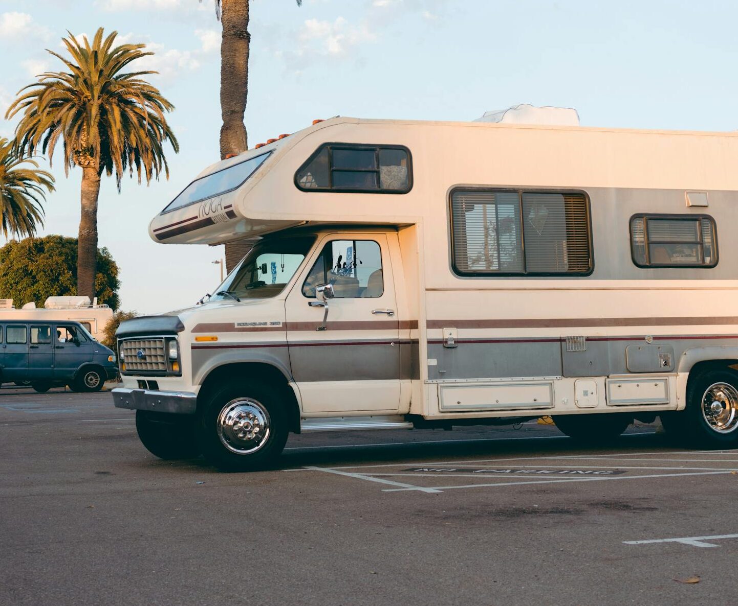 RVs parked under palm trees in a bright daylight urban parking lot setting.